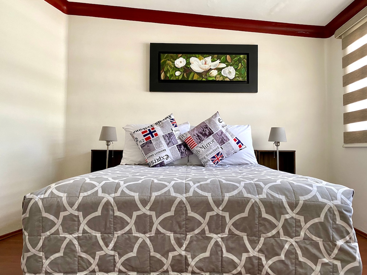 A cozy sleeping area features a spacious bed with a patterned gray blanket. Two small lamps sit on wooden nightstands flanking the bed. The walls are adorned with a floral artwork, complemented by natural light filtering through stylish blinds.
