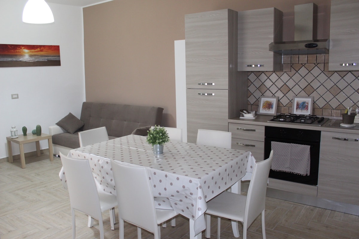 A modern kitchen and dining area is presented, featuring a large table surrounded by white chairs. A gray sofa is positioned nearby, and the kitchen includes light cabinetry and a gas stove. A decorative painting and a small plant add subtle touches to the space.