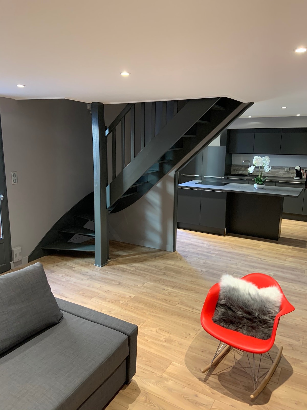 A modern interior features an open layout with a staircase leading to the upper level. The sleek kitchen is visible in the background, equipped with dark cabinetry and modern appliances. A gray sofa and a red chair with a fur throw add contrast to the warm wooden flooring.