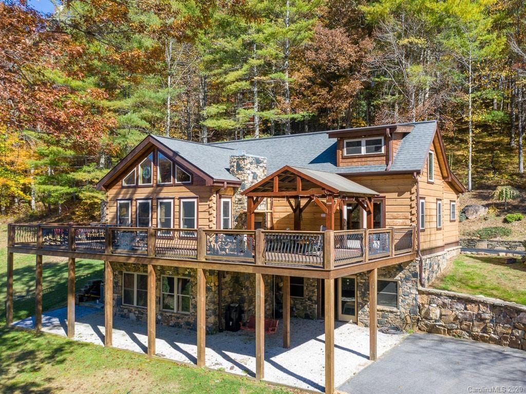 The cabin's exterior showcases a rustic design with a combination of wood and stone elements. A large deck extends around the front, providing ample outdoor space. Lush trees and vibrant foliage surround the property, highlighting its natural mountain setting.