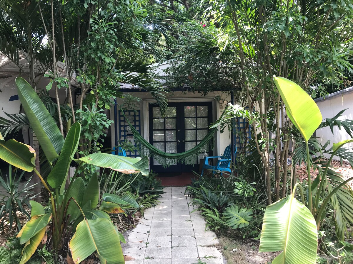 A pathway leads through lush greenery to the entrance of a private cottage. Tropical plants and trees frame the entrance, where a hammock is suspended between two wooden posts. Blue outdoor chairs are positioned nearby, evoking a serene outdoor space.