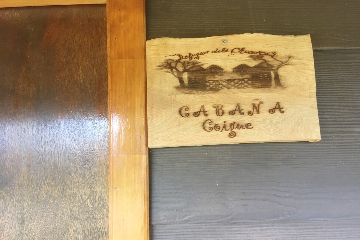 A wooden sign is displayed at the entrance, featuring the engraved text 'Cabaña Cotuye' alongside illustrations of rustic cabins and trees. The natural wood texture adds a warm touch to the inviting atmosphere.