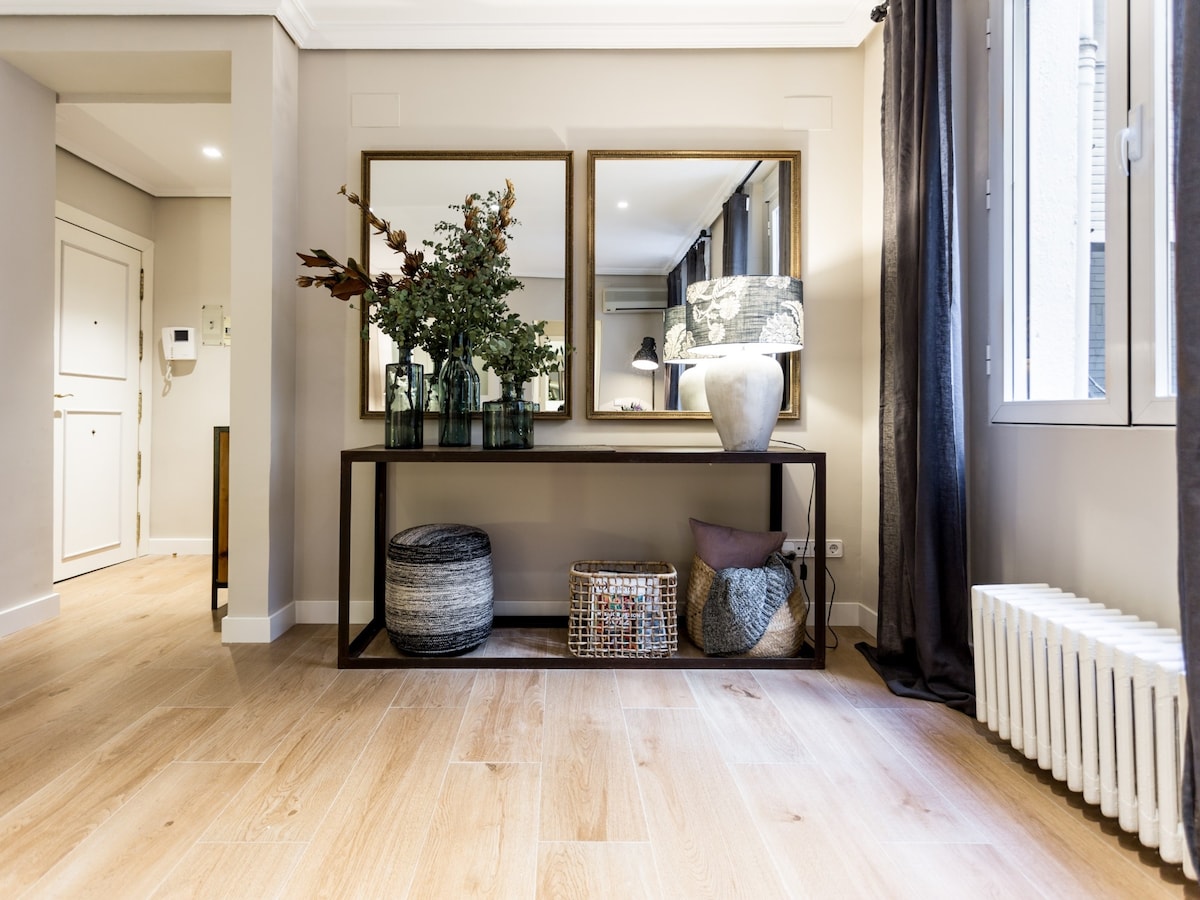 A welcoming entryway features a console table with decorative items, including vases and a stylish lamp. Two large mirrors reflect the space, enhancing the light from the windows. The flooring consists of light wood, while textured baskets add interest to the area.
