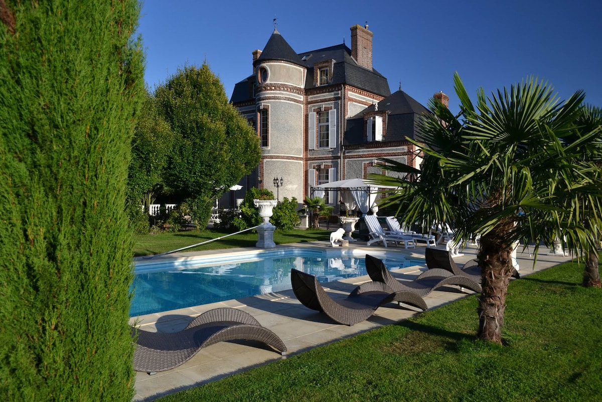 A charming 19th-century château is framed by lush greenery and trees, with a sparkling outdoor pool in the foreground. Comfortable lounge chairs are positioned around the pool, inviting relaxation under clear blue skies.