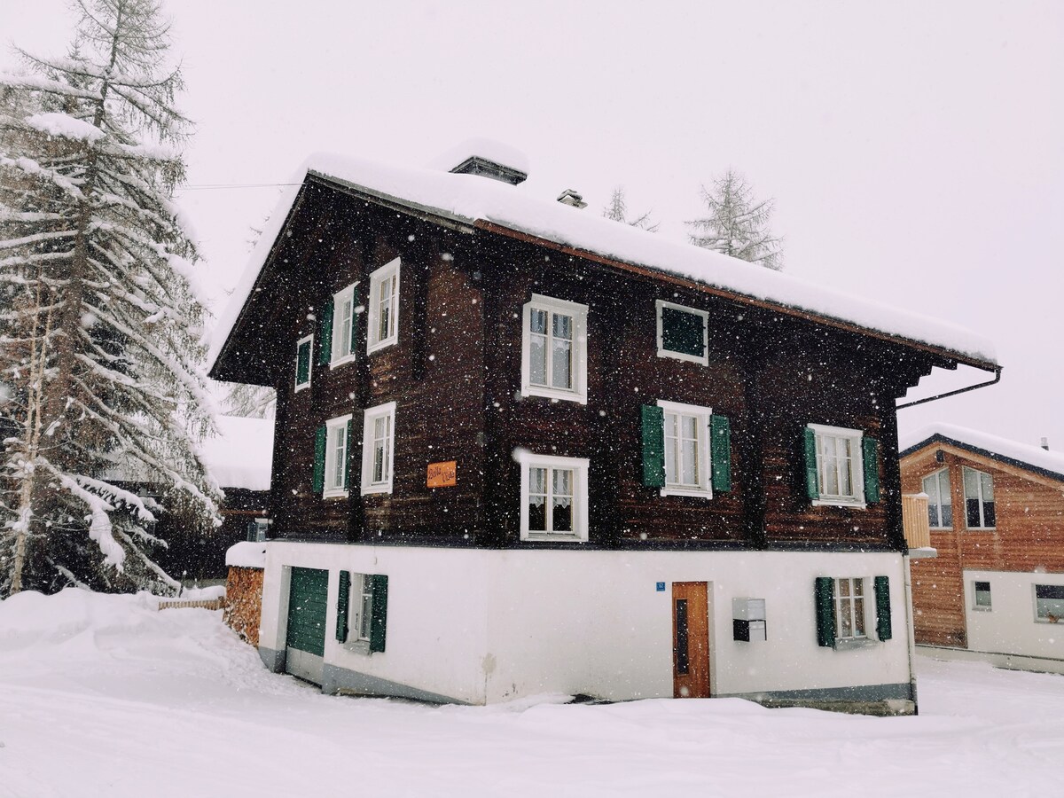 A traditional wooden house stands under a blanket of snow, with dark brown exterior walls and white accents. Green shutters adorn the windows, while snowflakes gently fall around it, creating a serene winter scene.