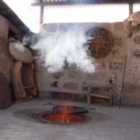 An indoor space is depicted, featuring a traditional stone oven with smoke rising from its circular opening. The rustic walls and wooden wheel decoration add character to the environment, while benches are positioned along the sides, providing a communal feel.
