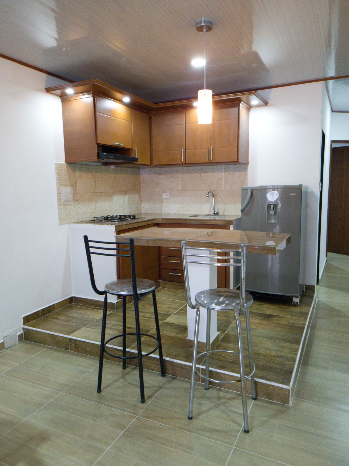 A compact kitchen space is highlighted by wooden cabinetry and modern appliances. A sleek refrigerator and stove are positioned alongside a stone countertop. Two high stools enhance the inviting atmosphere of the kitchen area, fostering a casual dining experience.