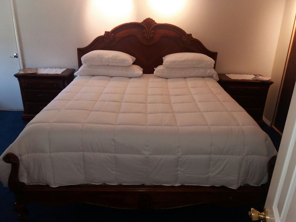 A spacious bed is positioned centrally in the room, adorned with a fresh white comforter and neatly arranged pillows. Dark wood nightstands flank each side, offering an elegant touch to the overall design.