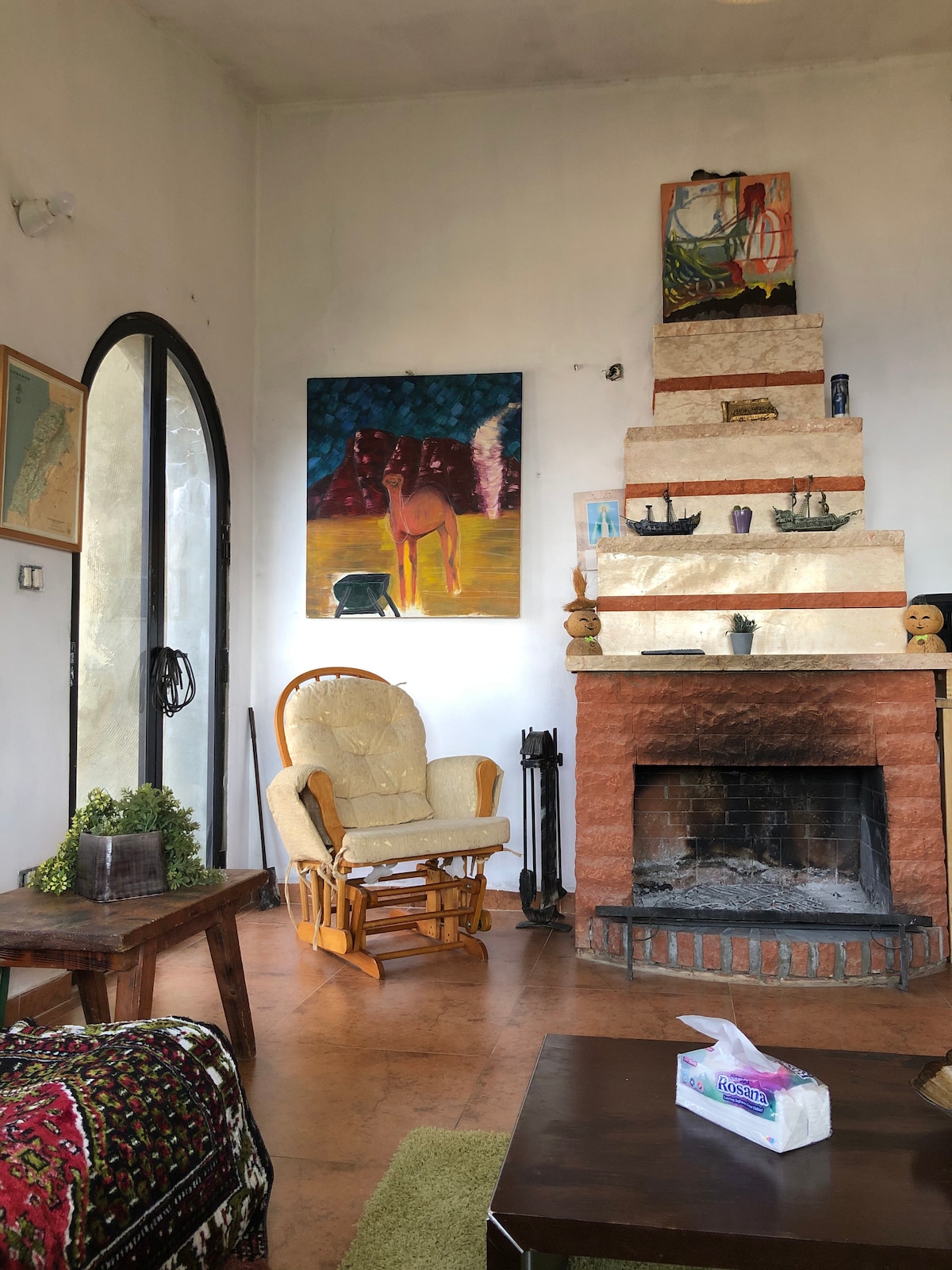 A cozy living area features a prominent fireplace, complemented by colorful artwork on the walls. A comfortable rocking chair is positioned near the hearth, inviting relaxation. Natural light enters through an arched window, illuminating the warm tones of the earthy floor.