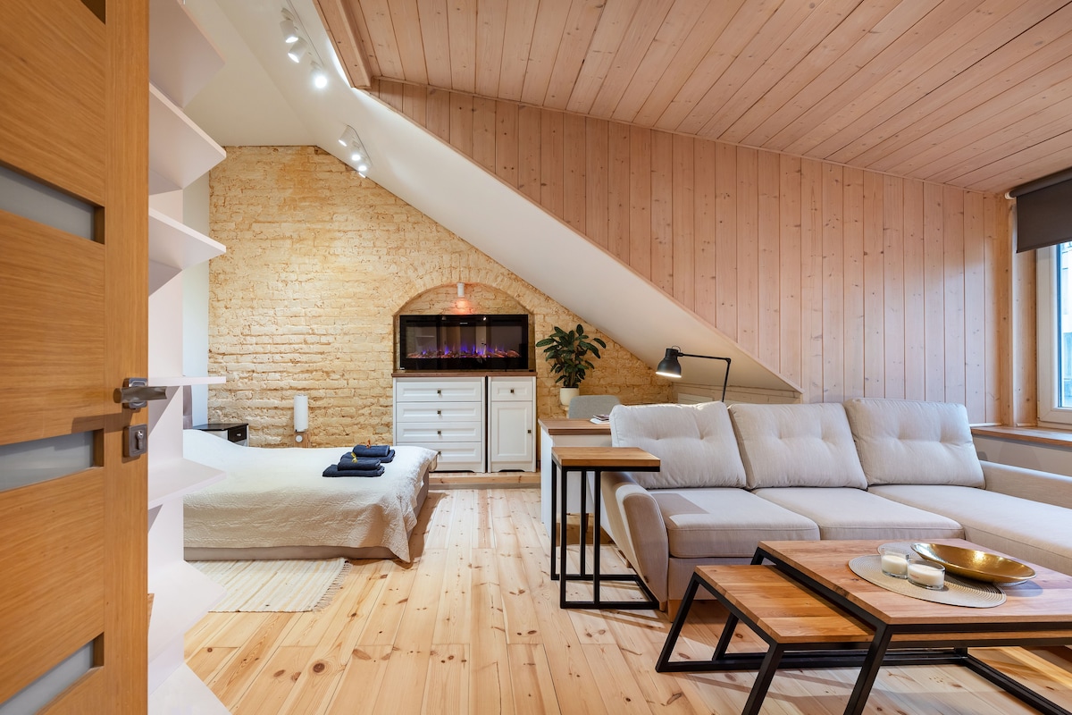 A cozy living area features a soft gray sectional sofa accompanied by wooden side tables. A modern fireplace is framed by exposed brick walls. The room showcases a sloped wooden ceiling, creating an inviting ambiance. A neatly made bed is positioned in the background.