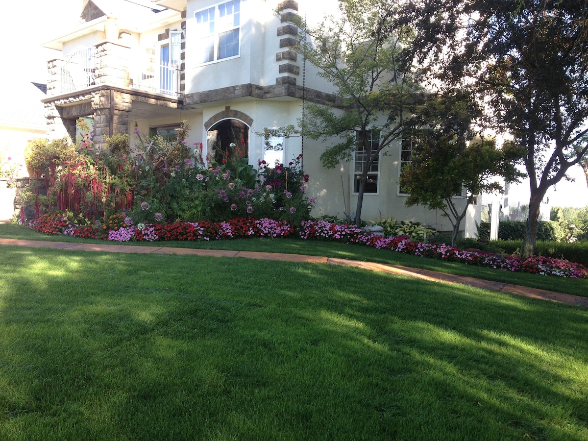 A well-maintained exterior showcases a garden adorned with vibrant flowers in various colors. Lush green grass complements the colorful blooms, while the structured facade of the home is highlighted by large windows and decorative stone accents.