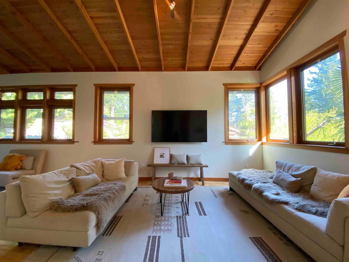 A sunlit living area features two modern sofas adorned with throws, positioned around a central coffee table. Large windows provide abundant natural light, showcasing the greenery outside. A mounted television is centrally located on the wall, enhancing the space's welcoming feel.