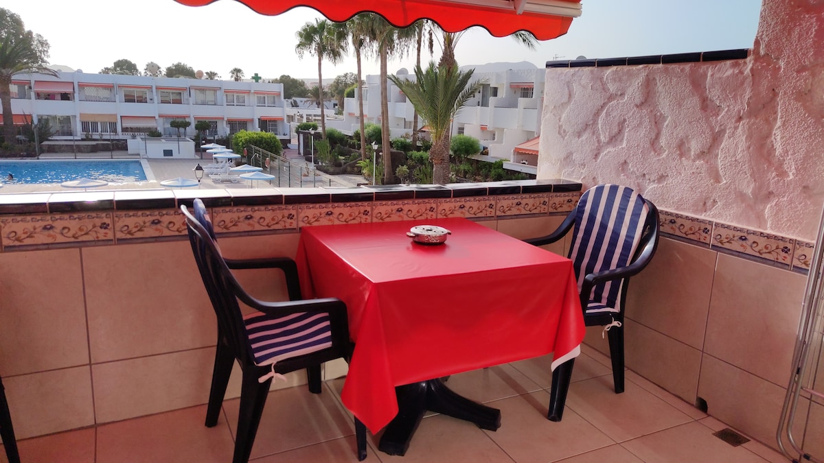 A private terrace is equipped with a red table and two black chairs. The area is shaded by an orange awning, providing a comfortable space to relax. In the background, a shared pool and palm trees can be seen, along with the surrounding accommodations.
