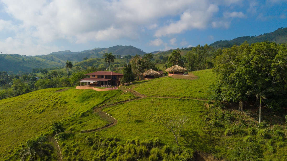 A spacious guesthouse is situated on a verdant hillside, surrounded by lush greenery and rolling mountains. The landscape features various trees and an outdoor seating area with thatched-roof structures, all under a partly cloudy blue sky.
