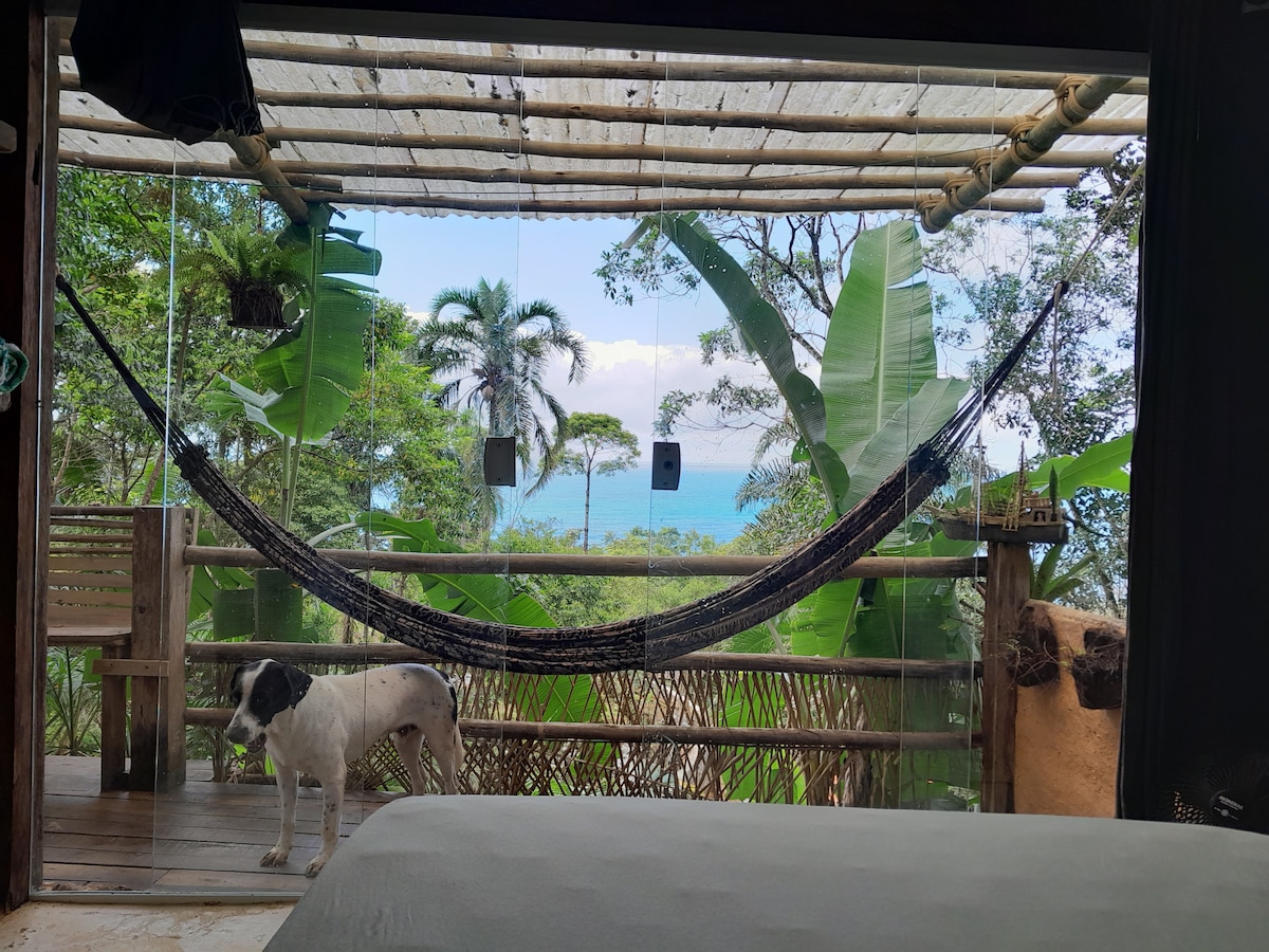 A glass-enclosed space features a hammock suspended between wooden posts, with a dog standing quietly nearby. Lush green foliage and palm trees frame a view of the ocean in the background, creating a serene environment that invites relaxation.