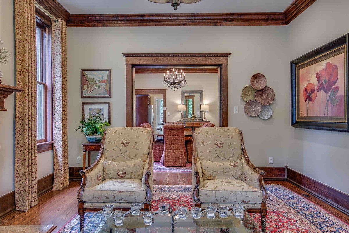 Two elegant armchairs with floral patterns are positioned on a patterned rug, inviting relaxation. Framed artworks adorn the walls, and a chandelier hangs from the ceiling, illuminating the cozy living space that connects to another room in the background.