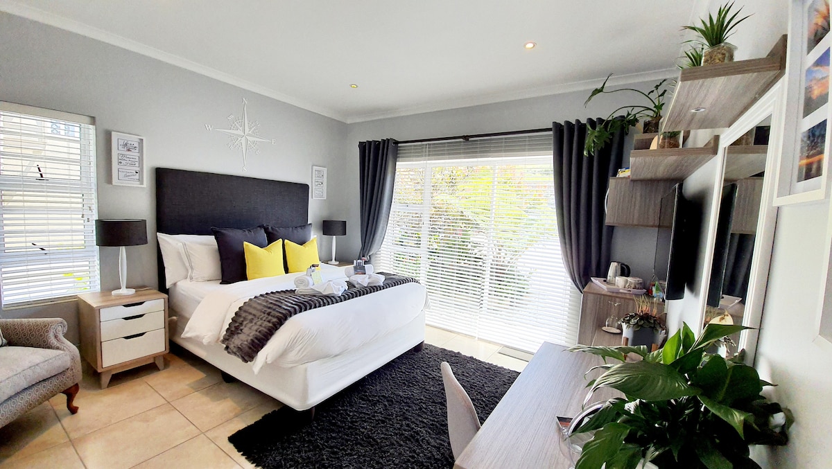 A spacious guest suite is described, featuring a queen size bed layered with soft bedding and decorative pillows. A modern work station is positioned nearby, with a comfortable chair. Natural light filters through large windows, complemented by sleek curtains, while greenery adds a touch of freshness.