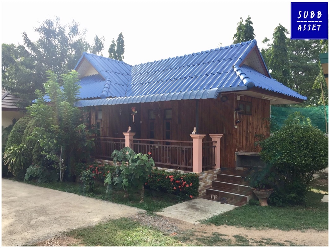 A wooden house with a blue roof is surrounded by lush greenery, including shrubs and trees. The front porch features decorative columns and steps leading to the entrance. Natural light reflects off the glossy blue tiles, enhancing the inviting exterior.
