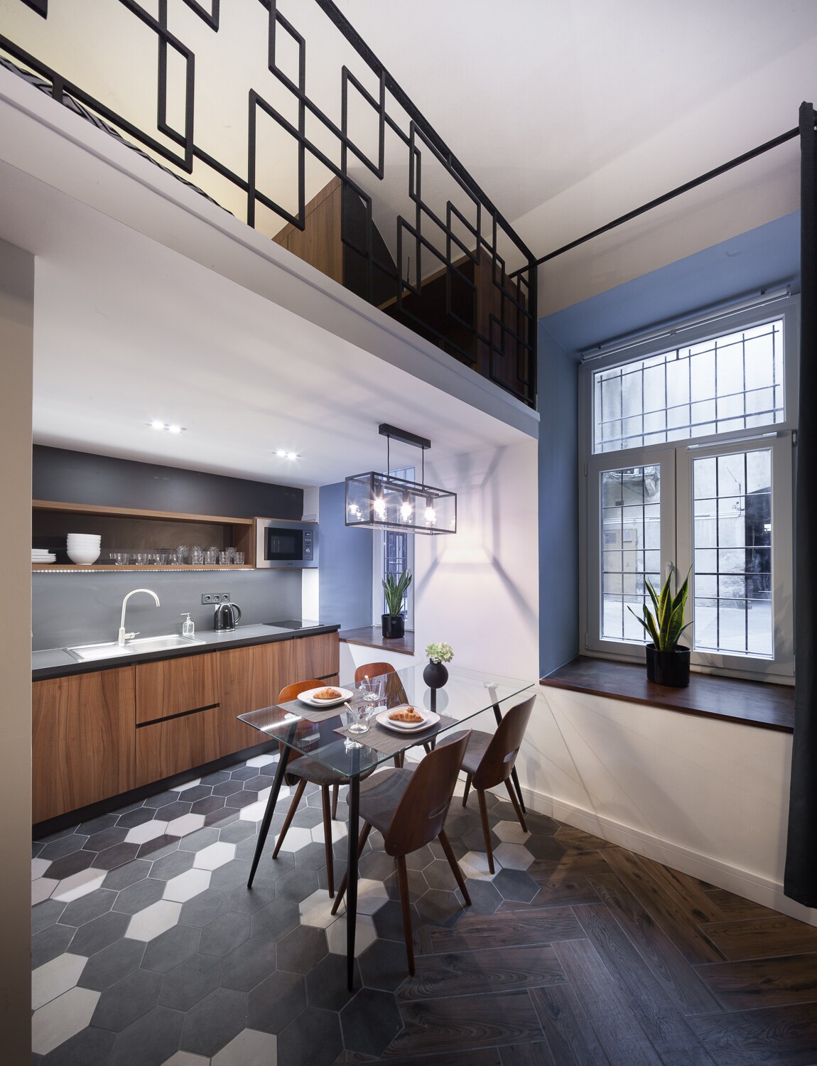 A modern kitchen and dining area feature sleek cabinetry in warm wood tones, with a dining table set for four. Natural light enters through a large window adorned with potted plants, casting shadows on the patterned floor tiles.