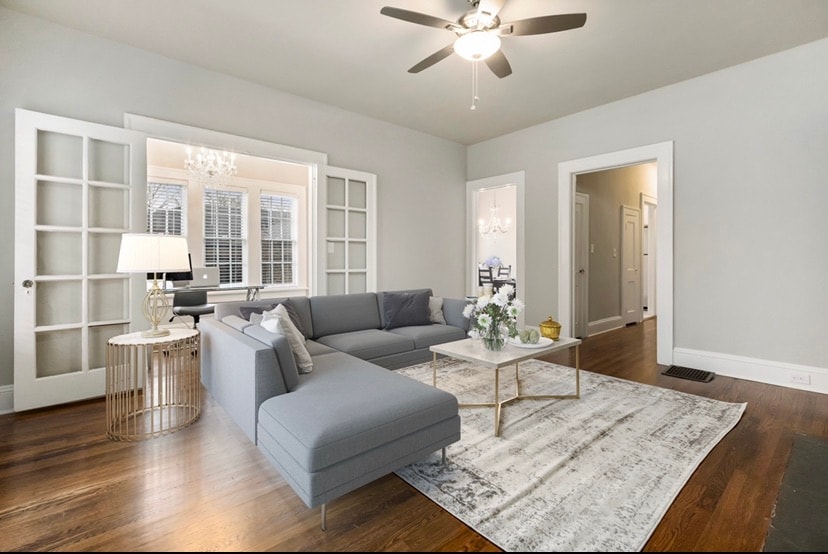 A spacious living area features a gray sectional sofa positioned around a low coffee table. A light rug lies beneath the seating arrangement, enhancing the hardwood floor. French doors lead to additional rooms, allowing natural light to illuminate the space.
