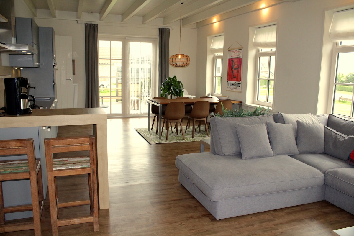 The spacious living area features a large sectional sofa facing a dining table surrounded by modern chairs. Natural light enters through multiple windows, and a woven pendant light adorns the ceiling. The hardwood floor complements the inviting atmosphere of the open layout.