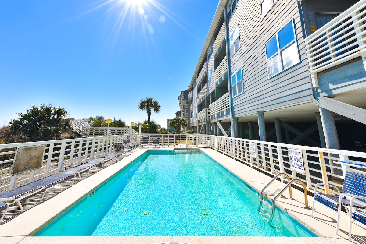 A swimming pool is surrounded by a spacious deck featuring lounge chairs. Sunlight reflects off the clear water, with palm trees and a sandy area visible in the background. The building structure has multiple floors with large windows overlooking the pool.