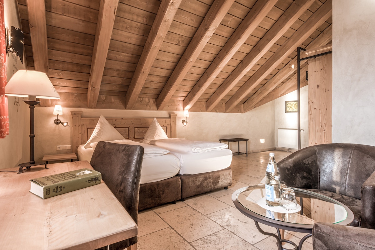 The room features a vaulted wooden ceiling, providing a sense of spaciousness. A cozy seating area includes dark upholstered chairs and a small round table. A large bed is styled with comfortable white bedding, complemented by decorative pillows. Soft lighting creates a warm ambiance.