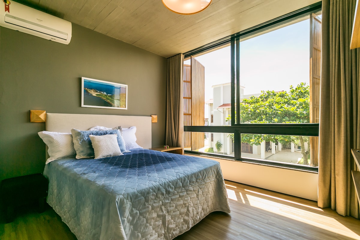 A spacious bedroom features a comfortable bed with light blue bedding. Natural light floods in through large windows, flanked by curtains. A small side table is positioned beside the bed, and artwork hangs on the wall, enhancing the serene atmosphere.
