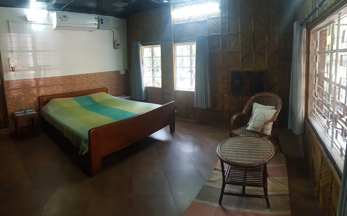 A spacious room features a wooden bed with a light green and blue blanket. Natural light enters through multiple windows with light curtains, illuminating the bamboo wall paneling. A comfortable wicker chair and small side table are positioned near the window.