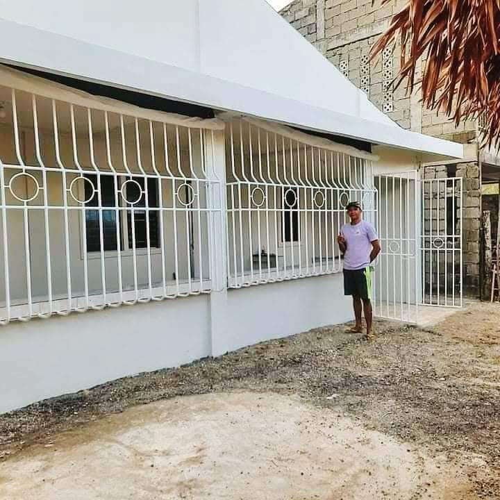 The exterior of the bungalow house is presented, showcasing a clean white facade with decorative railings. Large windows allow natural light to enter, while a sandy area surrounds the structure. The roof is sloped, complemented by a glimpse of tropical plants.