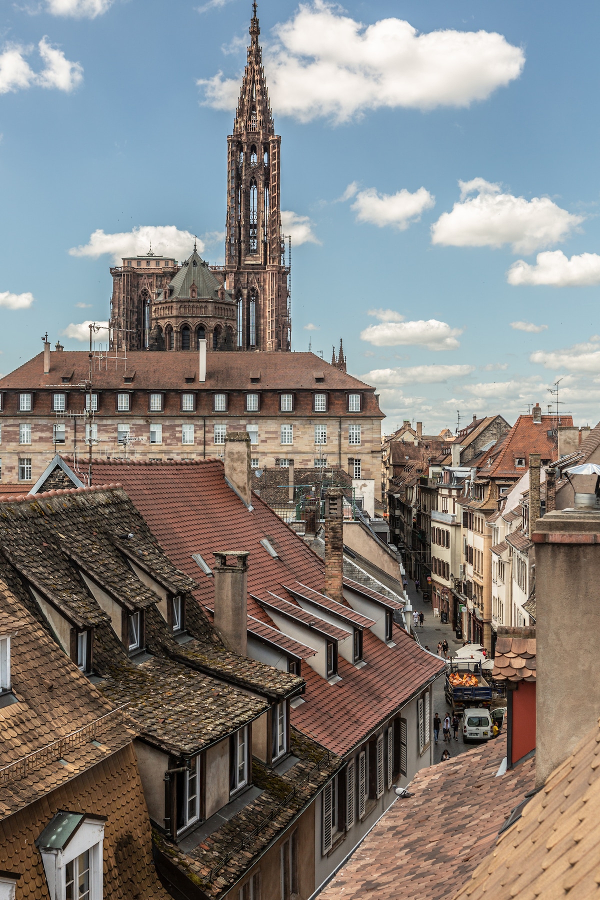 The image presents a view of the Strasbourg skyline, featuring the intricate architecture of the Strasbourg Cathedral rising above surrounding rooftops. A mix of historic buildings, with varied roof textures and colors, is visible in the foreground, set against a backdrop of clear blue sky and scattered clouds.