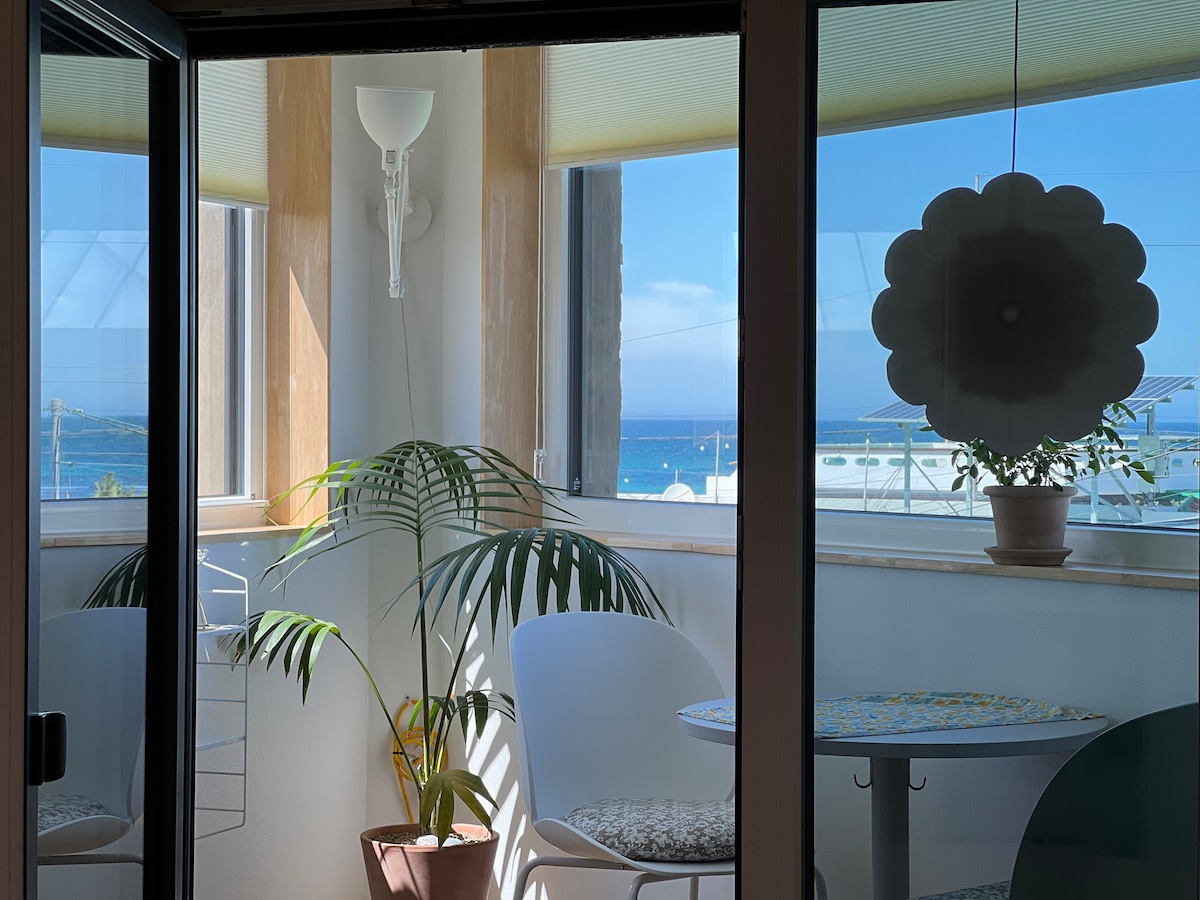 A sunlit dining area features a small table surrounded by white chairs, offering a view of the sea through expansive glass doors. A potted plant adds a touch of greenery, creating a bright and inviting nook for relaxation.