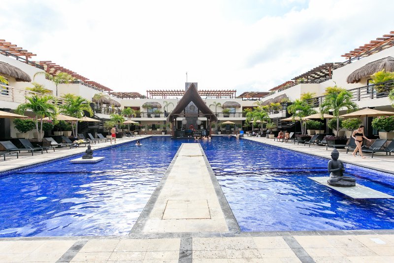 A large pool stretches across the center of the complex, surrounded by palm trees and shaded lounge chairs. Asian-inspired sculptures are placed at both ends of the pool, while thatched-roof structures offer additional shade. Guests can be seen enjoying the space under a clear sky.
