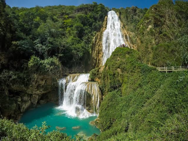 A cascading waterfall is set against a lush green hillside. Clear turquoise water collects at the base, surrounded by dense vegetation. The scene is framed by rocky cliffs, creating a serene natural environment.