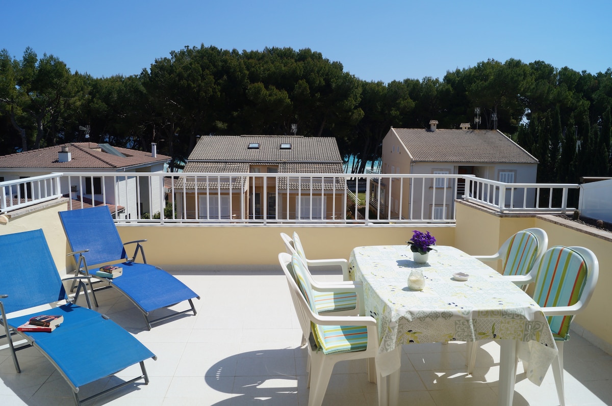 A spacious terrace is equipped with a table and four chairs, surrounded by two lounge chairs. Sunlight fills the area, with views of nearby greenery visible in the background. The peaceful setting suggests a comfortable outdoor space for relaxation and dining.