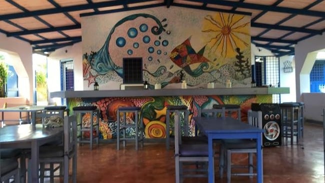 An inviting communal area features a vibrant mural depicting waves, fish, and sun. A long bar counter is complemented by several blue chairs and tables arranged for social interaction. Natural light enters through the open spaces, creating a cheerful environment.