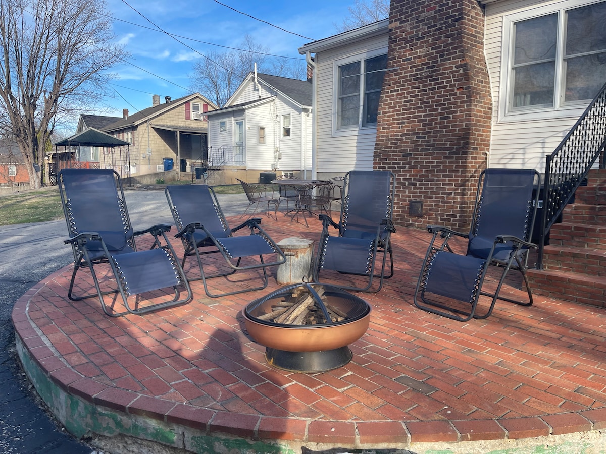 A circular brick patio area is shown, featuring four black lounge chairs positioned around a central fire pit. The area is surrounded by a grassy lawn, with additional buildings visible in the background.