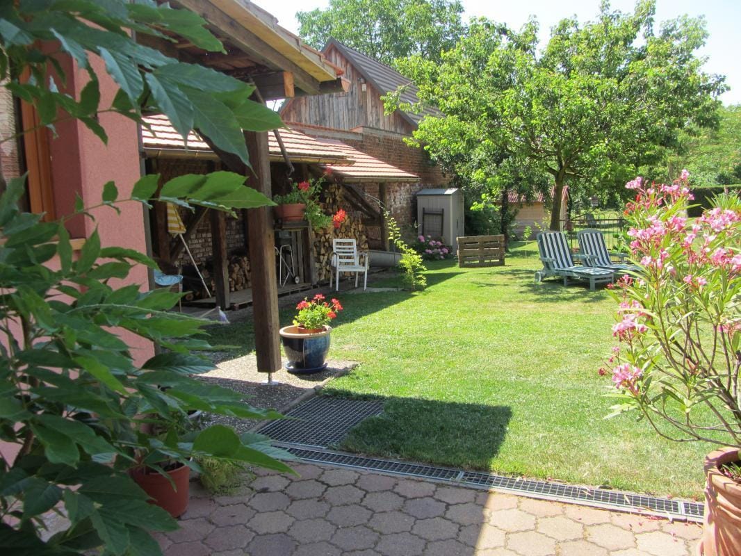 A spacious garden area features well-kept greenery, bordered by vibrant flowerpots. Sun loungers are arranged invitingly on the lawn, complemented by shade from surrounding trees. A shelter and garden furniture are visible, creating a functional outdoor space suitable for relaxation.
