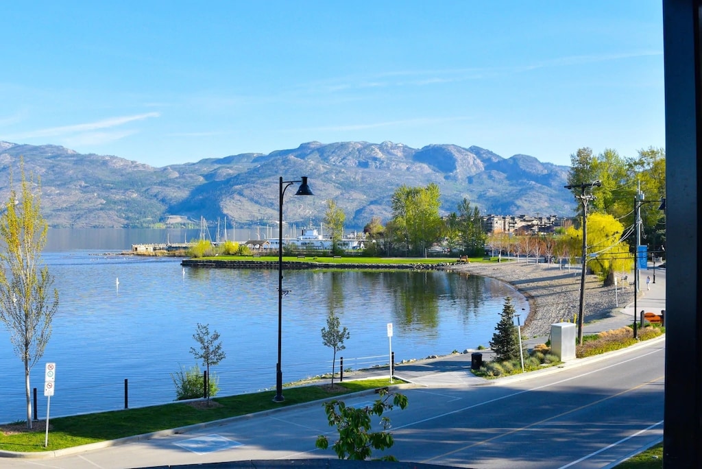 A serene lakeside view is captured, featuring calm waters reflecting the surrounding mountains. Along the shoreline, trees and grass areas create a natural landscape. A road runs parallel to the lake, lined with streetlamps and plantings, inviting peaceful strolls by the water.