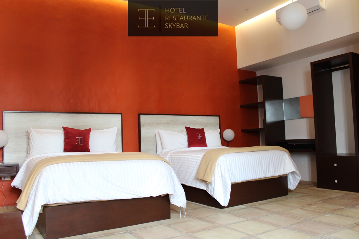 Two spacious beds are arranged in a room with an orange accent wall. Soft lighting is provided by bedside lamps, and a minimalist desk with a mirror is visible. A wardrobe is positioned against the wall, contributing to the functional layout of the space.