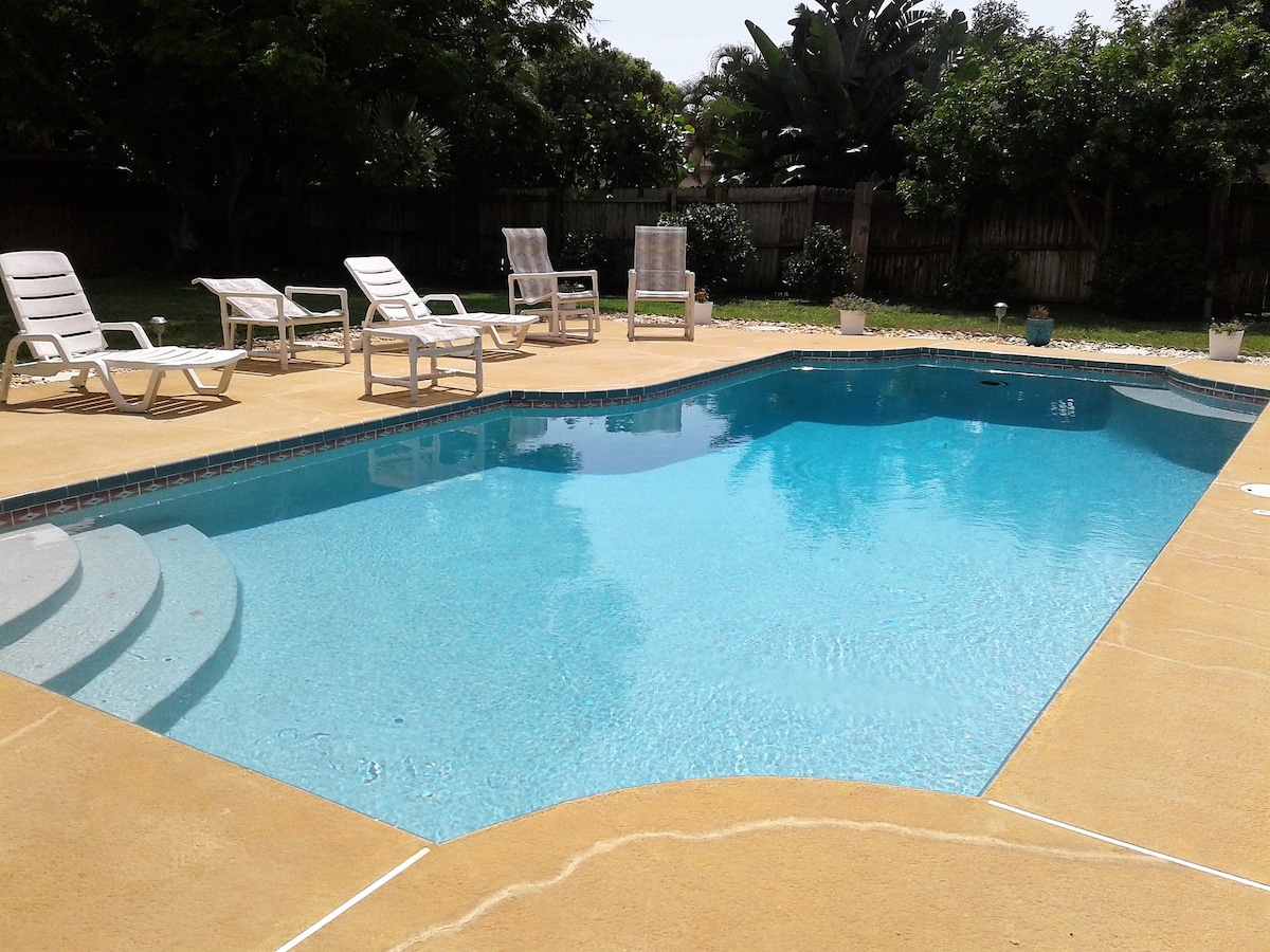 A spacious pool area is surrounded by a paved deck, featuring a clear blue swimming pool with gradual steps. Several lounge chairs are positioned nearby, along with potted plants adding a touch of greenery to the outdoor space.