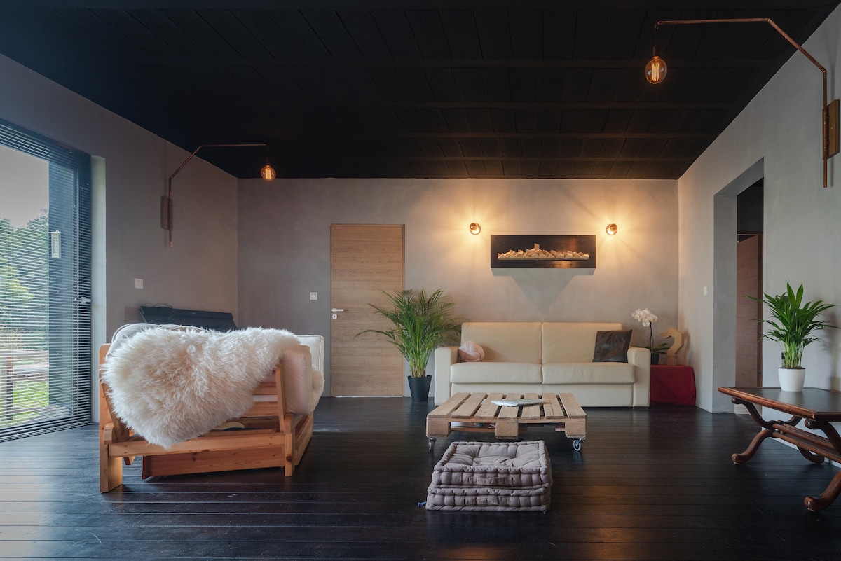 A modern living area is furnished with a white sofa, a wooden daybed, and a rustic coffee table on wheels. Ambient lighting is provided by wall-mounted fixtures, while large glass doors open to the outside. Plant accents enhance the natural feel of the space.