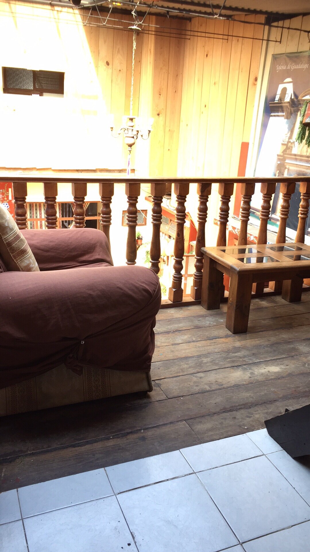 A cozy balcony area features a wooden railing and a small wooden table. A comfortable chair, draped with a soft cover, invites relaxation. The flooring is a mix of polished wood and tile, with sunlight filtering through an opening above.