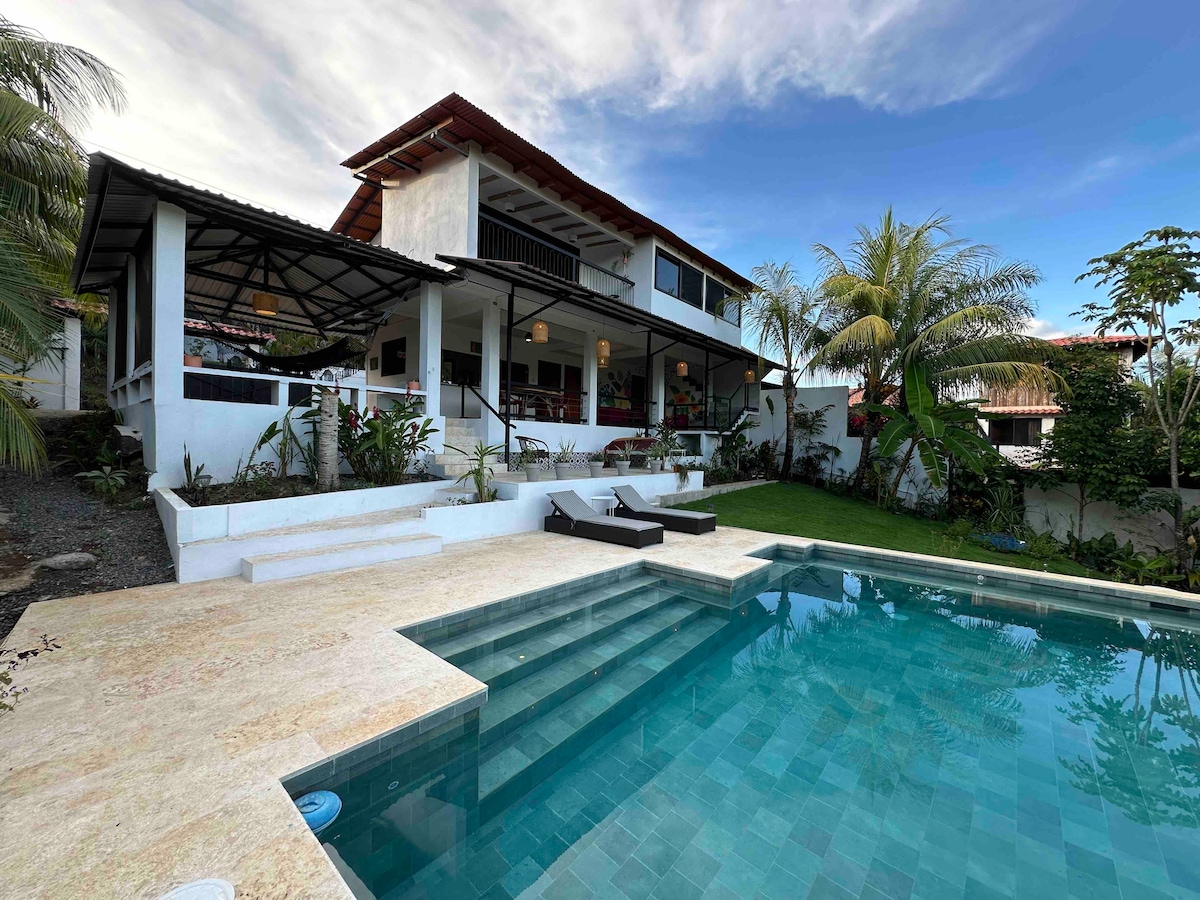 The exterior of a two-story house is highlighted, showcasing a private Balinese-style pool and lounging area. Lush greenery surrounds the property, with steps leading to the pool from a spacious deck. Large windows offer views into the home's inviting interior.
