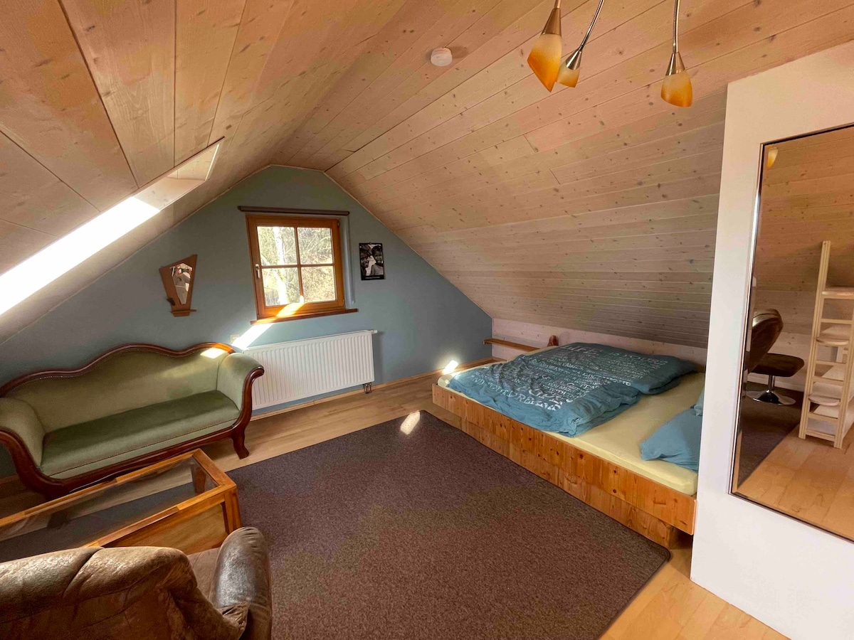 A loft-style room features a spacious bed with blue bedding, complemented by a wooden frame. A green seating area includes a vintage sofa, and natural light enters through a slanted roof window. A cozy carpet adds warmth to the wooden floor.