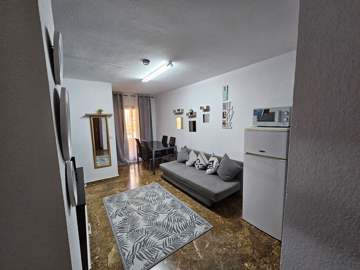 A spacious living area is visible, featuring a comfortable gray sofa bed and a large rug with a leaf pattern. A dining table with four chairs is arranged nearby. Light enters through large windows, and decorative elements are displayed on the walls.