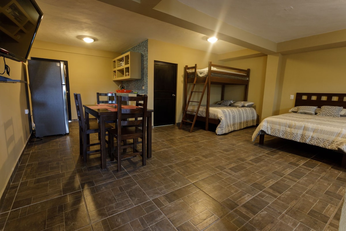 A spacious room features a dining table with six chairs and a kitchen area visible in the background. Two sets of beds, including a bunk bed, occupy one side of the room, while a television is mounted on the wall opposite the beds.