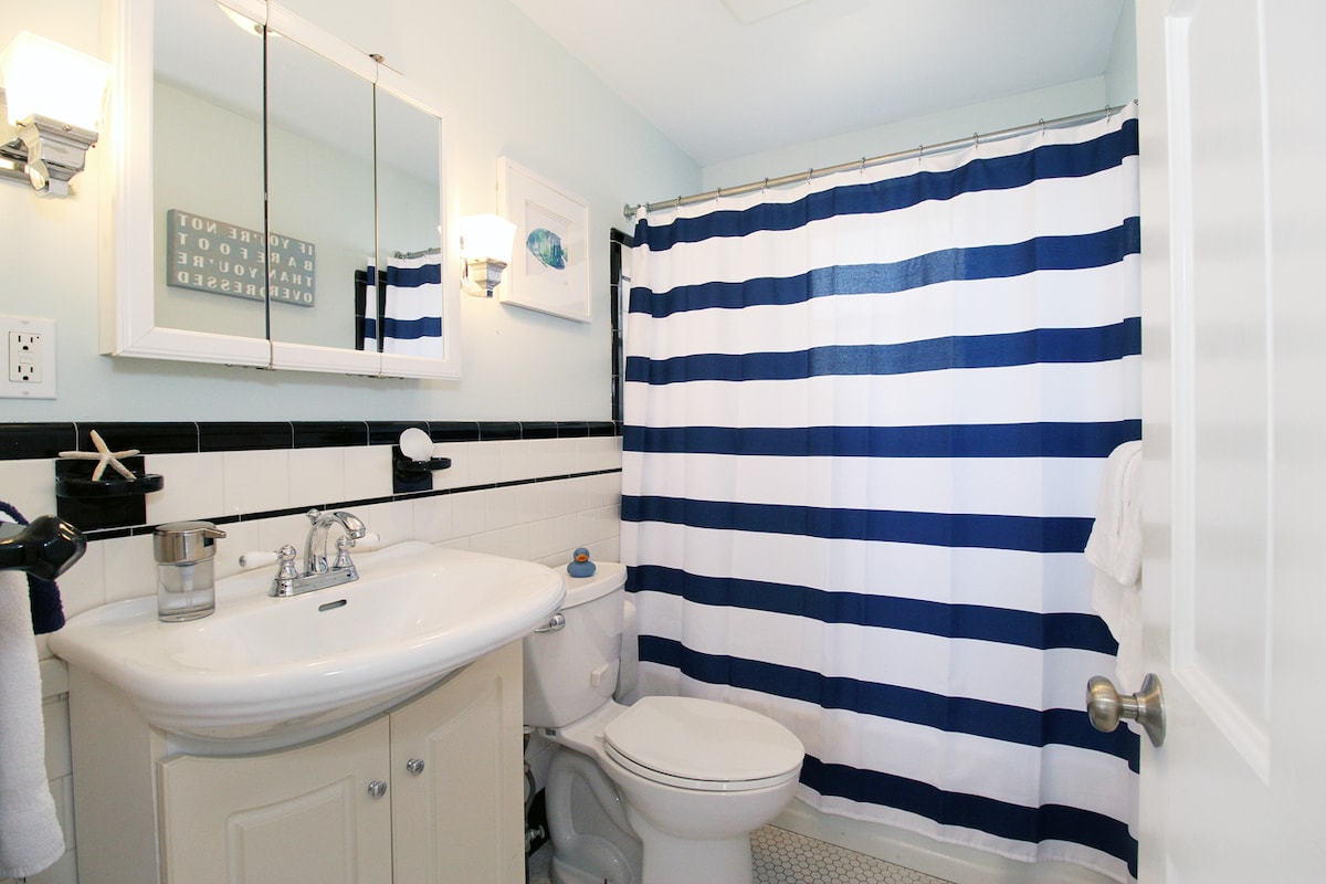 A well-lit bathroom features a white sink and toilet, accompanied by a striped shower curtain in navy and white. A framed piece of art adorns the wall, and a mirror reflects the light from the wall sconces, enhancing the clean aesthetic.