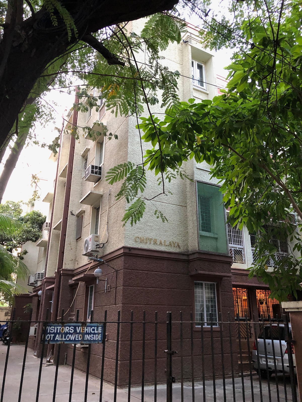 A multi-story building is framed by lush green trees, showcasing a beige exterior with a contrasting green section. Windows are visible on multiple levels, emphasizing the structure's height. A gated entrance is indicated by a sign stating 'Visitors Vehicle Not Allowed Inside.'