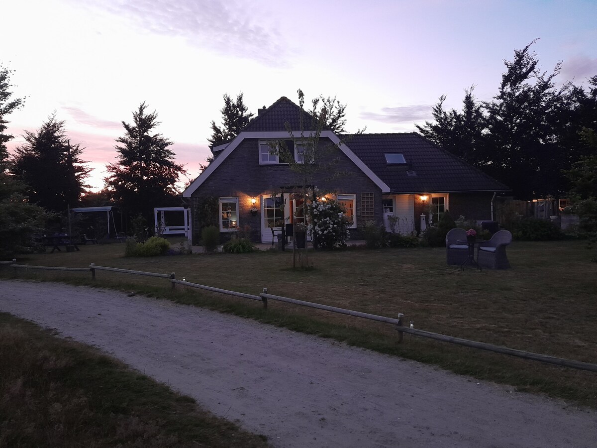 A charming house is set against a twilight sky, surrounded by greenery. Pathways lead toward the home, framed by trees, with outdoor seating visible on the lawn. Soft lighting illuminates the exterior, creating a calming atmosphere in the evening.